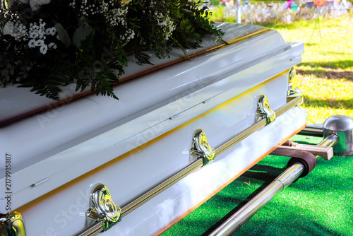 Coffin sits on green grass at burial site, flowers decorations surround cemetery area, family stands nearby.