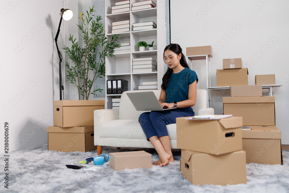 Fototapeta premium Asian woman entrepreneur working on laptop, managing online e commerce business from home, surrounded by shipping boxes for order fulfillment and small business growth