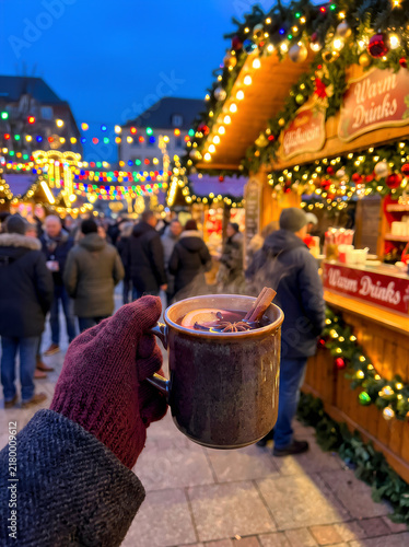 Hand in wool glove holding mug of hot mulled wine at festive outdoor Christmas market with lights and wooden fair stalls decor. Merry Christmas and New Year family celebration concept with copy space.