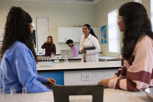 Indian instructor teaching teenage students in lab coat holding tablet in school lab with flasks