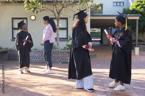 Diverse graduates chatting in courtyard wearing gowns holding diplomas one wearing pink cardigan