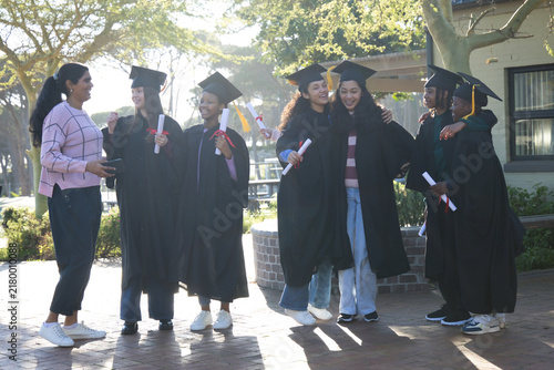 Diverse group of seven youth graduates in black gowns caps posing on courtyard, holding diplomas