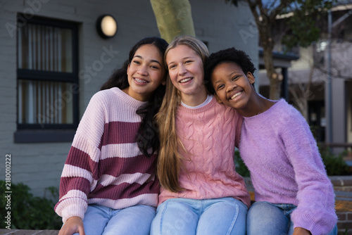 Diverse teenage females sitting close on low wall bench by tree trunk in courtyard wearing sweaters