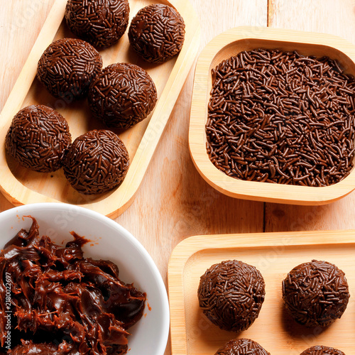 Classic Brazilian chocolate brigadeiro finished with sprinkles, served on a bamboo tray as part of a culinary arrangement on a wooden table.