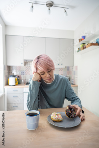 Obraz na płótnie Mature woman sits stressed beside colorful donuts, struggling with PMS cravings and sugar addiction while trying to resist pastries, reduce sugar consumption and regain dietary self-control