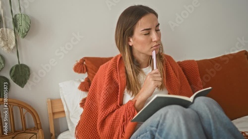 Young woman writing notes in a notebook on a bed wrapped in an orange blanket inside a building with pillow and rattan chair; quiet contemplation.