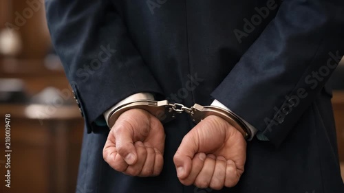 Close up of arrested man in formal suit standing with hands handcuffed behind back during court trial, white collar crime, suspect in handcuffs, legal sentence, justice concept shot in courtroom