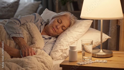 Elderly woman sleeping peacefully in cozy bed under lamp light with medicine bottle pills and glass of water on bedside table, senior illness treatment, healthcare, home care concept shot