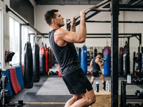 Strong male MMA fighter doing pull-ups on a metal bar during training in a boxing gym with punching bags