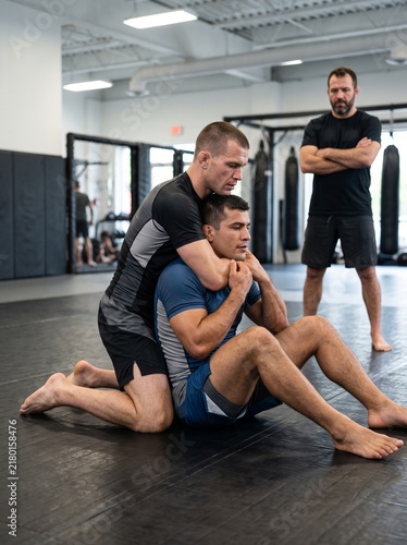 MMA fighters practicing a rear naked choke submission hold with a coach watching in a gym