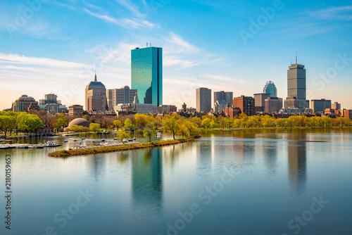 A vibrant daytime view of the Boston skyline featuring John Hancock Tower and Prudential Center, with reflections in calm waters of the Charles River.