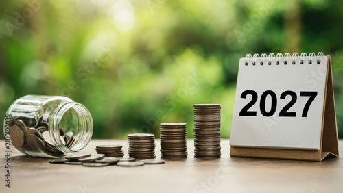 A desk calendar for 2027 stands next to stacked coins and a glass jar on a wooden surface. Concept for financial planning and future investment.