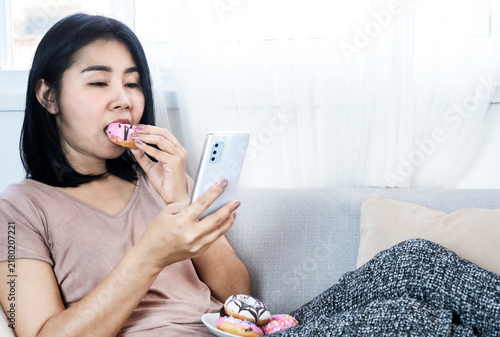 Obraz na płótnie Asian woman eating a pink donut while using a smartphone on sofa at home