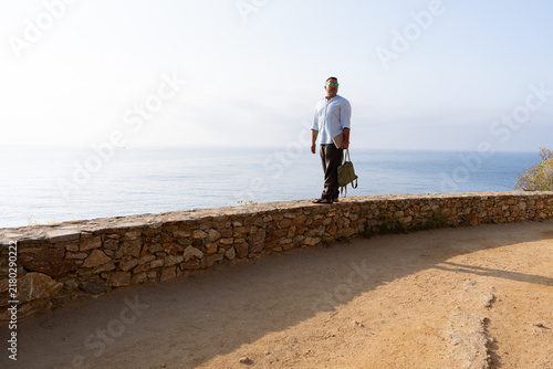 Obraz na plátně Man with backpack and laptop standing on stone wall contemplating ocean horizon