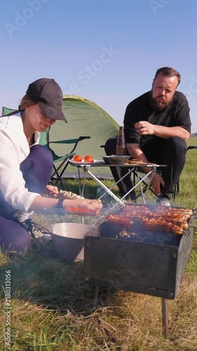 Young couple enjoying a sunny day outdoors, grilling sausages and shashlik skewers on a barbecue. Friends having a picnic with a tent set up in a beautiful green meadow on a summer day
