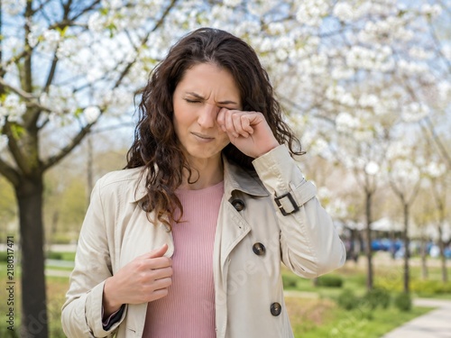 Woman suffering from allergy rubbing itchy eye near blooming tree in spring park. Seasonal allergic rhinitis caused by flower pollen. Hay fever symptoms and pollen allergy treatment concept