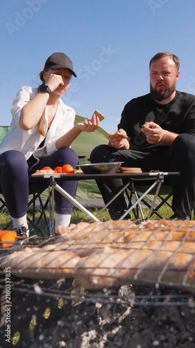 Young couple sitting in camping chairs by their tent, enjoying freshly grilled meat from the barbecue and having a conversation during their outdoor adventure on a sunny day in nature