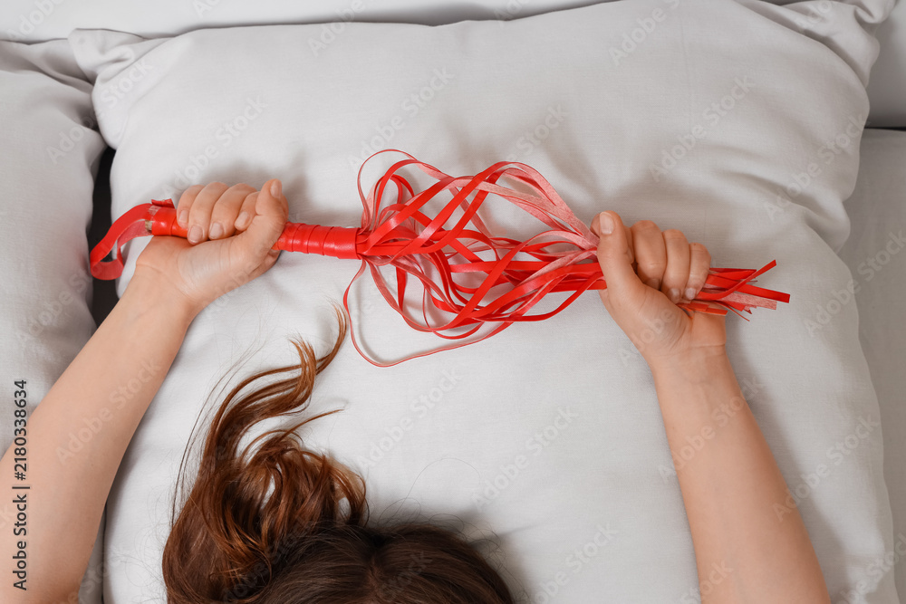 Fototapeta premium Bdsm lifestyle and intimacy, woman holding a red leather flogger while lying on a white bed, of adult play and sexual exploration in bedroom