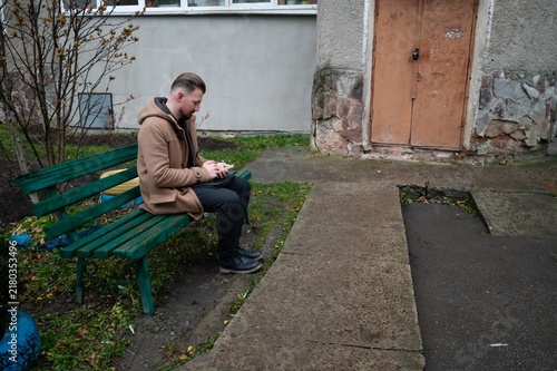 Quadro su tela Young man sitting on a green bench absorbed in his smartphone in an urban enviro