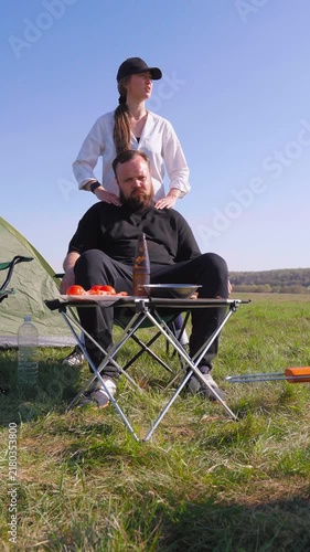 Young caring wife giving a relaxing shoulder and neck massage to her tired bearded husband during a picnic while camping together in a green field on a sunny day with a tent in the background