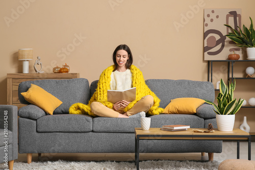 Beautiful young woman with warm yellow plaid reading book while sitting on so...