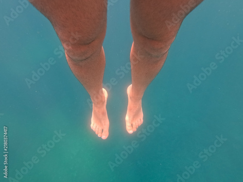 Canvastavla POV shot of bare male legs and feet floating vertically downwards in the clear, turquoise waters of the Mediterranean Sea