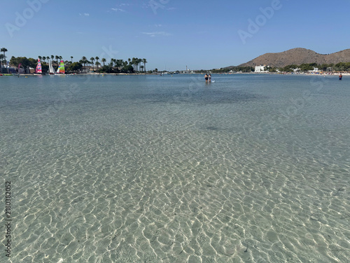 Fotografie Clear shallow water at Port d'Alcúdia beach, in Alcúdia, on the island of Mallorca, Balearic Islands, Spain, with rippled sand beneath the surface and the shoreline in the distance