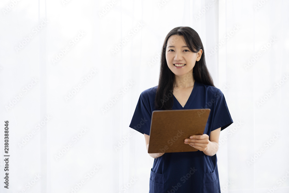Obraz premium Young Japanese female doctor in scrubs holding a clipboard and smiling at the camera by a bright window