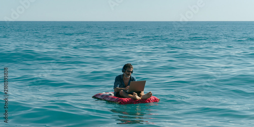 A man working on a laptop while floating on a raft in the middle of the ocean, enjoying the tranquil blue water and clear sky.