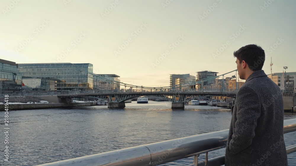 Obraz premium Man Gazing at Dublin Skyline at Dusk, Reflective Moment by the River Liffey, Contemporary Urban Scene.