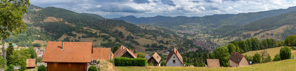 Obraz premium Munster, France - 08 20 2026: Panoramic view of houses on the mountain with the Munster Valley and the city in the High Vosges massif with forests and cloudy sky behind