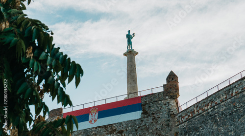 Canvastavla Pobednik monument and Serbian flag are framed by foliage above Belgrade Fortress