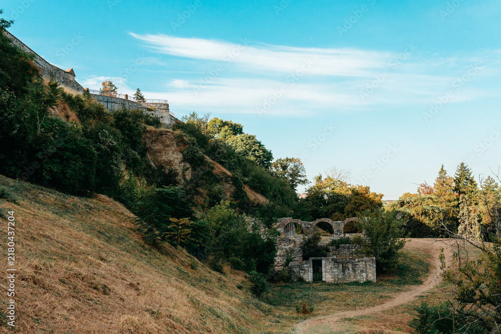 Fototapeta premium Stone ruins and old fortress walls stand on a grassy hillside in Kalemegdan, Belgrade. Archaeology, heritage tourism and historic travel concept in Serbia.