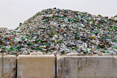 Huge pile of discarded glass bottles and broken containers behind concrete barriers at recycling facility, showing waste accumulation and material recovery