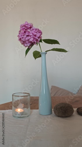 Beautiful pink hydrangea stem in a tall pastel blue vase placed next to a burning tea candle in glass and smooth zen stones on a tabletop against a light neutral background.