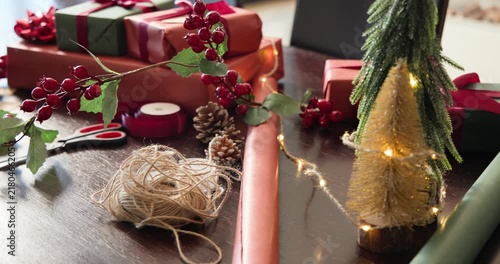 Camera pulling back, revealing twine, holly, red paper on dining table to show wrapping station