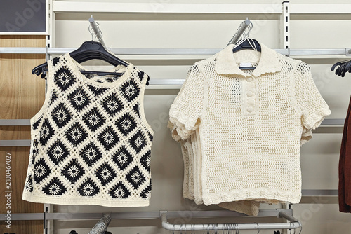 Crochet sleeveless top and cream knit polo shirt hanging on retail display rack, textured handmade fashion in neutral store interior