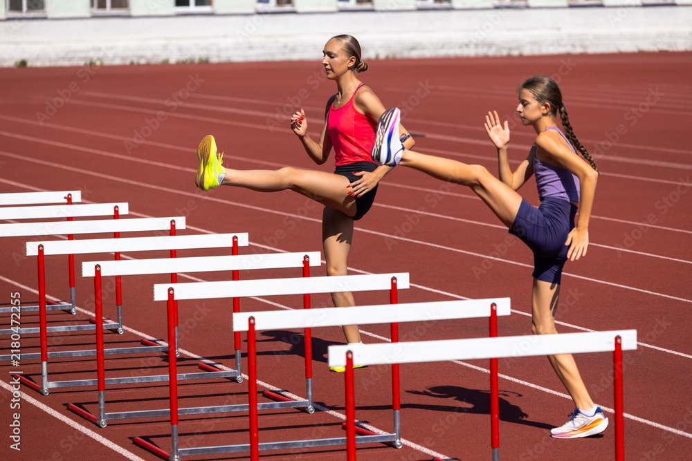 Fototapeta premium Adult and teenage female athletes lift legs over training hurdles. Concept of flexibility, dynamic stretching, coordination, high jump preparation and joint practice.