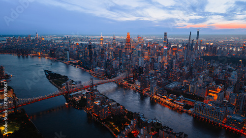 Twilight view of Midtown Manhattan skyline and Queensboro Bridge on East River. Illuminated skyscrapers and bridge reflect orange lights into dark river water.