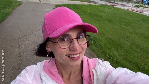 Close up pov portrait of a cheerful young woman wearing round eyeglasses and a pink cap smiling looking at camera while walking outdoors in a park.