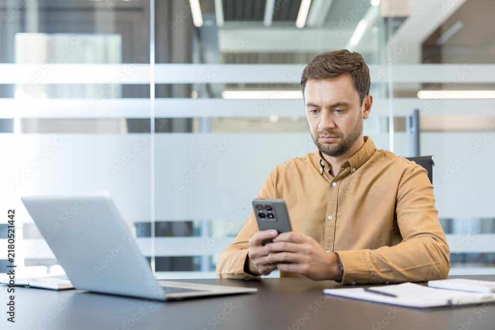 Fototapeta premium Businessman concentrating on his smartphone while sitting at a sleek desk with a laptop and documents, engaging with digital communication and technology in a corporate environment