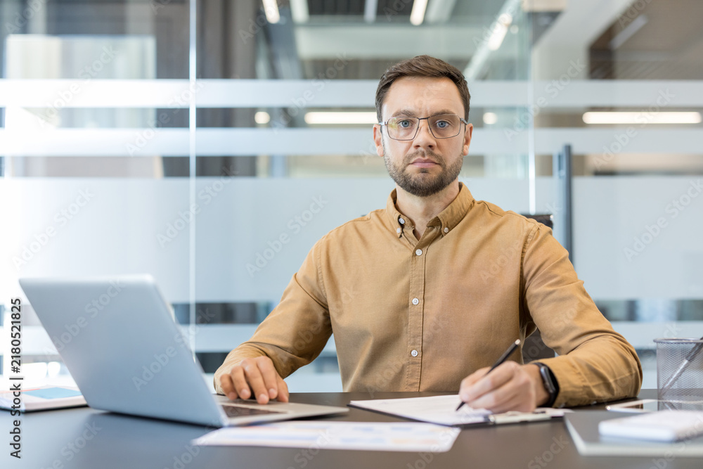 Obraz premium Confident businessman working diligently at a modern office desk, wearing glasses and a casual shirt, while typing on a laptop and taking important notes on paper