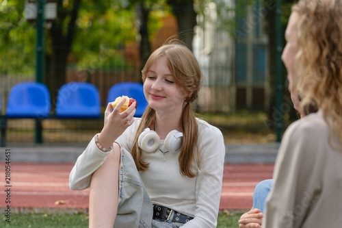 Teenage high school girl smiles while eating a red apple during a break on an outdoor school sports field. She sits on artificial grass with friends nearby and wears white headphones around her neck.