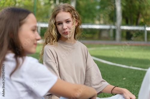 Quadro su tela Teenage high school girl with curly hair talks with a classmate while sitting on an outdoor school sports field