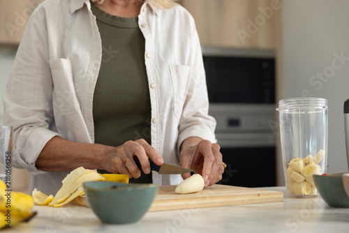 Senior woman trimming banana on cutting board and filling blender cup in kitchen wearing olive top