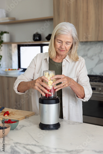 Senior woman preparing blended drink at modern kitchen counter with blender cup and fruit bowls