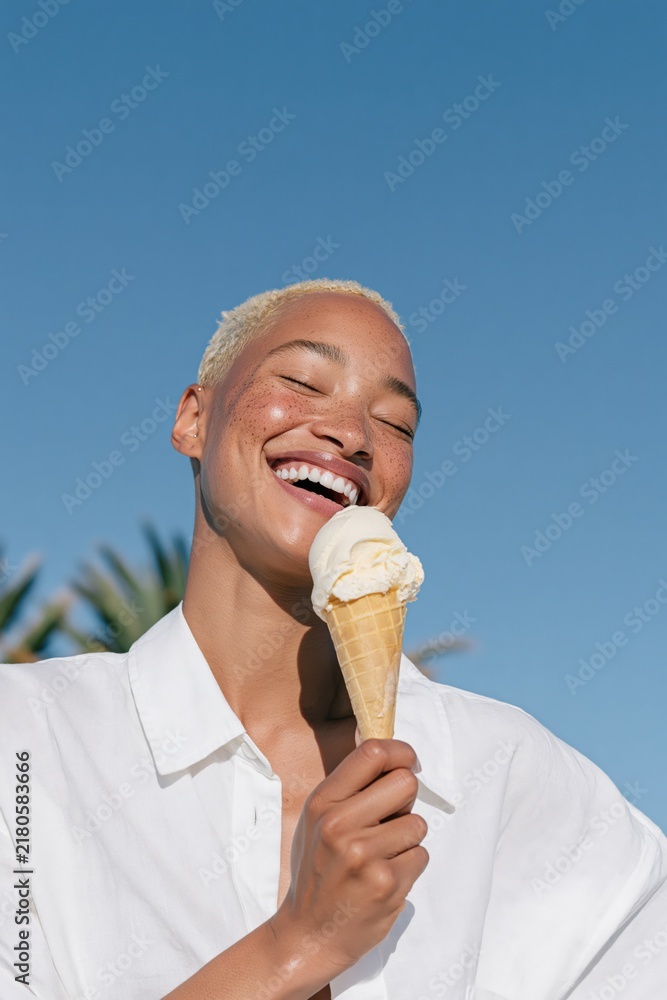 Obraz premium Joyful woman eating ice cream cone outdoors against blue sky for summer lifestyle banner and social media background use