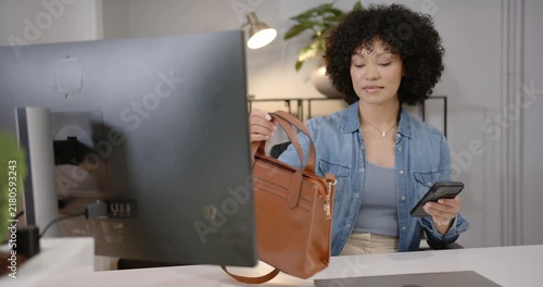 African American woman glancing at smartphone, placing brown handbag at desk and typing messages
