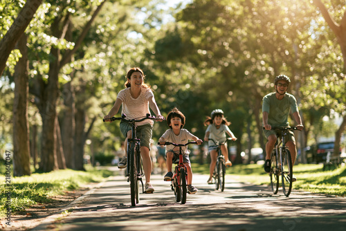 Happy Asian family cycling together outdoors, enjoying active lifestyle in sunny park with green trees, wearing casual clothes and helmets, bonding and smiling on summer day