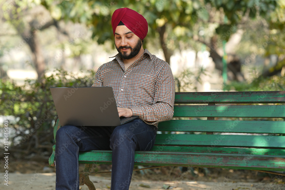 Fototapeta premium Young Indian Sikh man working on laptop while sitting on bench at summer park.